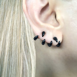 May include: A close-up of a person's ear with multiple black claw-shaped earrings. The earrings are arranged in a staggered pattern, with some pointing upwards and others pointing downwards.