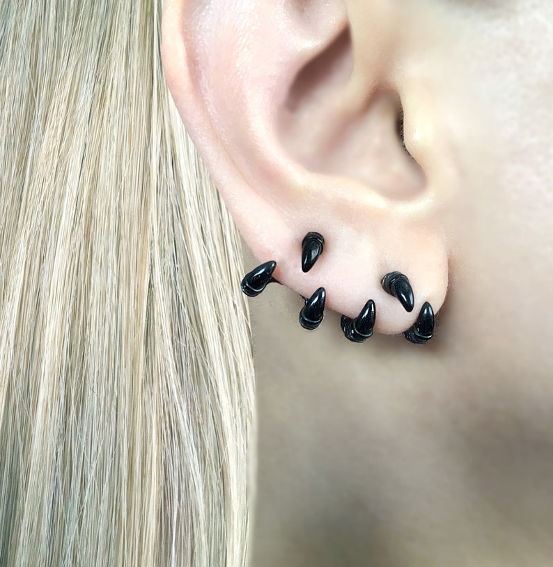 May include: A close-up of a person's ear with multiple black claw-shaped earrings. The earrings are arranged in a staggered pattern, with some pointing upwards and others pointing downwards.