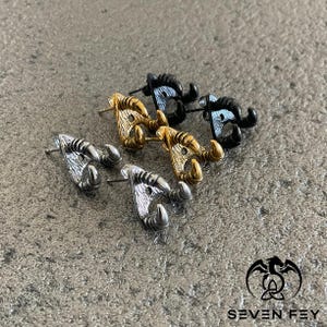 May include: Three pairs of earrings with a claw design. The earrings are made of metal and come in silver, gold, and black.
