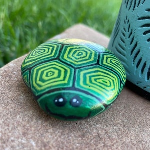 May include: A hand-painted green and yellow turtle rock with black eyes and a smile. The rock is sitting on a brown surface.