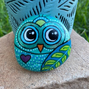 May include: Hand-painted rock art depicting an owl with large eyes, a purple heart, and green leaves on a blue background.