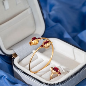 May include: A white jewelry box with a zipper closure, containing a gold bracelet and ring with red gemstone pomegranate designs.