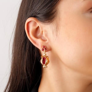 May include: Gold earrings with a pomegranate design. The earrings are adorned with red gemstones.