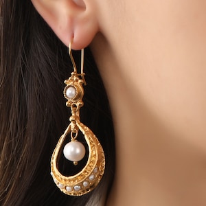 May include: Gold dangle earrings with a pearlescent drop and a smaller pearl at the top. The earrings have a delicate, ornate design.