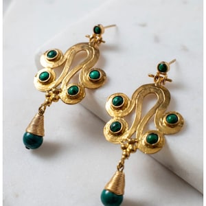 May include: A pair of gold-toned earrings with a unique, ornate design. Each earring features a series of circular green stones and a dangling teardrop-shaped green bead. The earrings have a post-back closure and are displayed on a white surface.