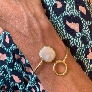 Puede incluir: Una pulsera dorada con una piedra grande, redonda y blanca, similar a una perla, y un peque&ntilde;o dise&ntilde;o de c&iacute;rculo abierto. La pulsera se lleva en la mu&ntilde;eca, con una tela estampada en el fondo.