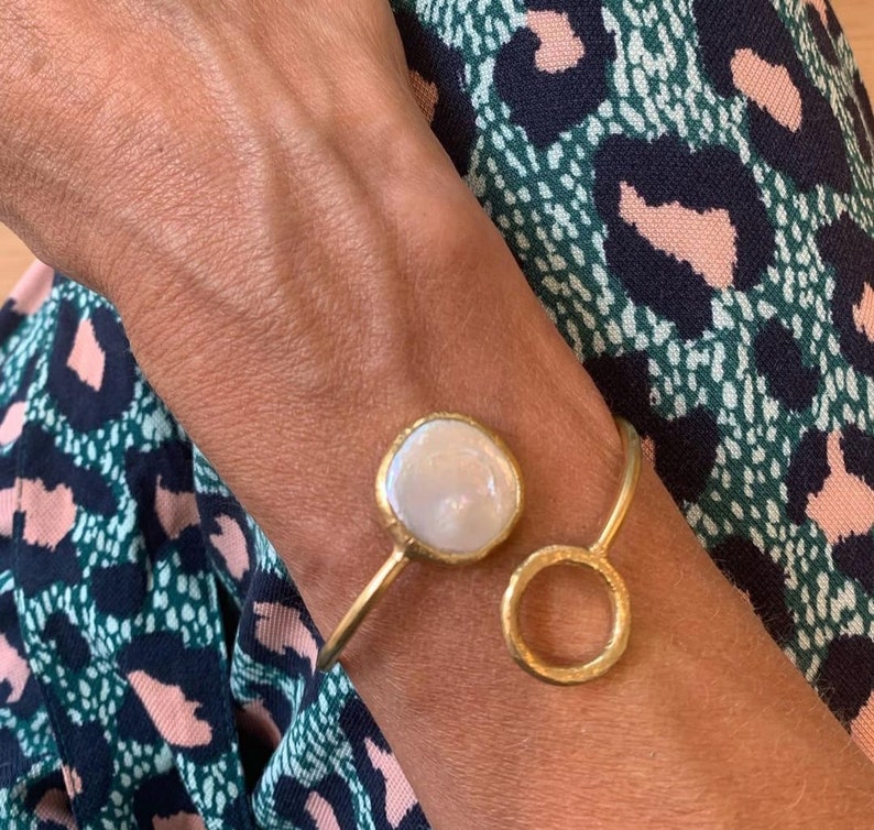 Puede incluir: Una pulsera dorada con una piedra grande, redonda y blanca, similar a una perla, y un peque&ntilde;o dise&ntilde;o de c&iacute;rculo abierto. La pulsera se lleva en la mu&ntilde;eca, con una tela estampada en el fondo.