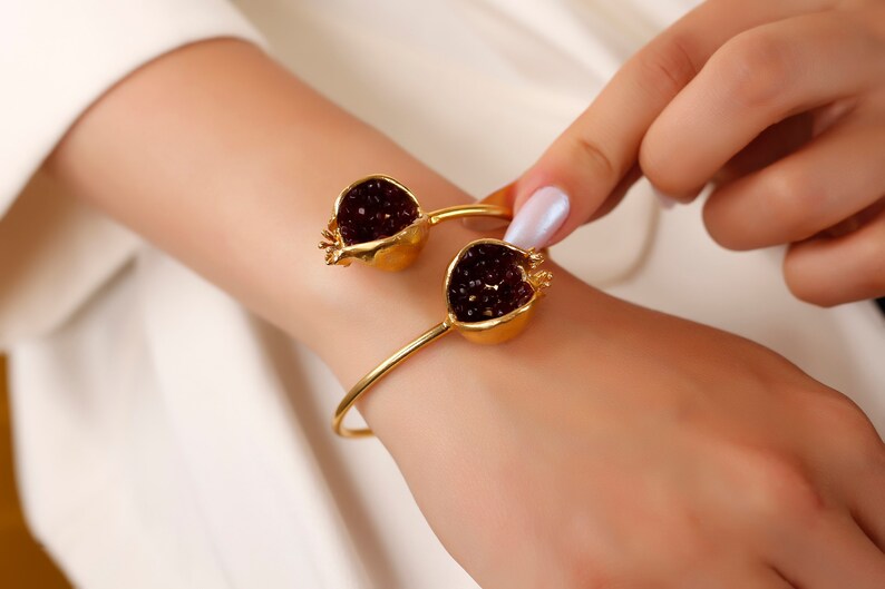 Puede incluir: Una pulsera de oro con dos abalorios en forma de granada. Los abalorios est&aacute;n llenos de cristales rojo oscuro.