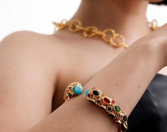 Puede incluir: Un brazalete de oro con piedras preciosas ovaladas de colores. El brazalete está abierto y tiene una bisagra en un lado.