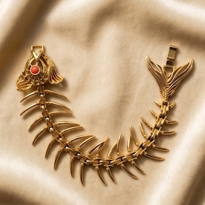 May include: A gold-toned metal bracelet shaped like a fish skeleton. The head features a small, round, orange-red cabochon stone as an eye. The body consists of articulated, curved rib-like segments, and the tail forms the clasp mechanism.