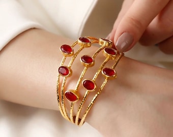 Puede incluir: Pulsera de brazalete de oro con múltiples bandas y acentos de piedras preciosas rojas.