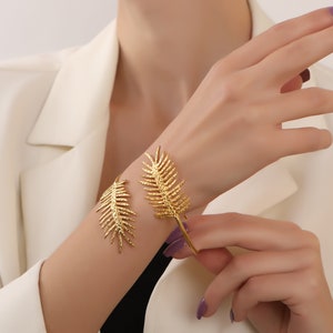 May include: A pair of gold leaf-shaped bracelets on a person's wrist. The bracelets are open and have a textured surface.