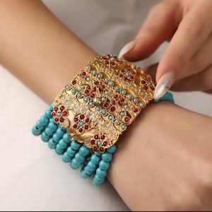 May include: A gold bracelet with a large, ornate, rectangular charm featuring intricate floral designs and red gemstones. The bracelet is made of turquoise beads.