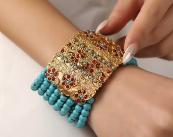 Puede incluir: Un brazalete de oro con un gran dije rectangular adornado con intrincados diseños florales y piedras preciosas rojas. El brazalete está hecho de cuentas turquesa.