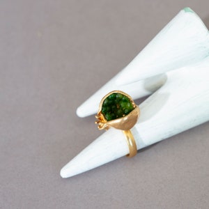 Può includere: Un anello d'oro a forma di melograno con pietre preziose verdi all'interno. L'anello è esposto su due porta anelli conici bianchi.