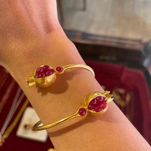 May include: A gold bangle bracelet with two pomegranate-shaped charms. Each charm is filled with red ruby-like stones.