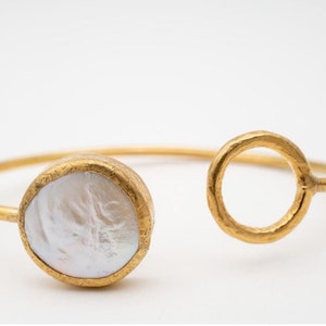 Puede incluir: Brazalete abierto dorado con una piedra grande, redonda y blanca con aspecto de perla en un extremo y un dise&ntilde;o circular en el otro. El brazalete tiene un acabado texturizado y un dise&ntilde;o minimalista.