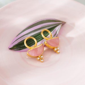 May include: A pair of gold earrings with pink stone accents and pearl details. The earrings are on a pink plate with a green leaf.