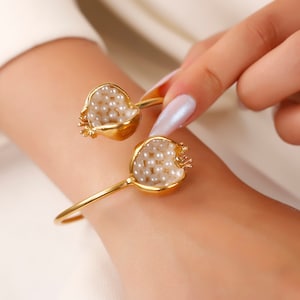 Puede incluir: Pulsera de oro con dos abalorios en forma de granada, cada uno lleno de perlas blancas.