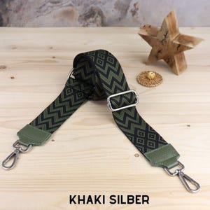 Peut inclure: Une sangle réglable à motif vert et noir avec des accessoires argentés et le texte "KHAKI SILBER".