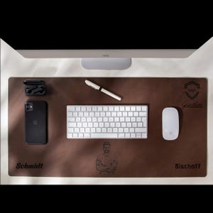 Könnte beinhalten: Eine braune Leder-Schreibtischunterlage mit einem personalisierten Design. Die Unterlage zeigt einen Namen, ein Logo und ein Porträt. Die Unterlage enthält außerdem eine Tastatur, eine Maus, einen Stift und ein Paar drahtlose Ohrhörer.