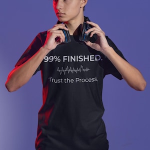 May include: Black t-shirt with the text "99% FINISHED." and "Trust the Process." in white. The shirt also features a sound wave graphic. The person in the image is wearing headphones.