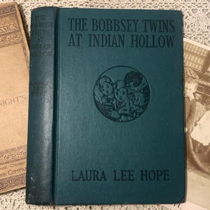 May include: A teal-colored book titled "The Bobbsey Twins at Indian Hollow" by Laura Lee Hope. The cover features an illustration of children. Beside it is a vintage book and a sepia-toned photograph.