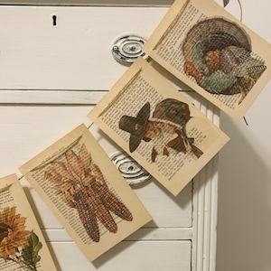 May include: Thanksgiving-themed garland featuring vintage book pages with images of a turkey, corn, and a pilgrim. The garland is strung on twine and hangs against a white dresser.