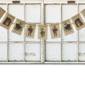 Puede incluir: Una festiva guirnalda navide&ntilde;a colgada frente a una ventana blanca. La guirnalda presenta tarjetas rectangulares de estilo vintage con ilustraciones navide&ntilde;as, incluyendo a Pap&aacute; Noel, un trineo y arreglos florales. Las tarjetas est&aacute;n impresas en papel envejecido.