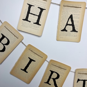 Pu&ograve; includere: Uno striscione fatto di vecchie pagine di libri con la parola "HAPPY BIRTHDAY" stampata in lettere nere. Lo striscione &egrave; legato insieme con uno spago.