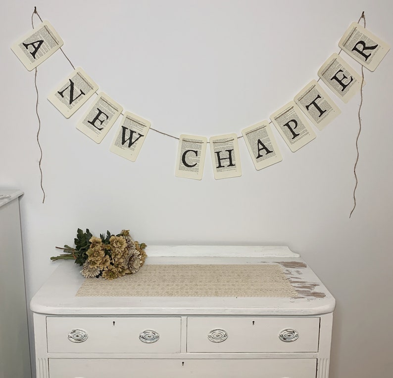 Pu&ograve; includere: Una cassettiera bianca con tre cassetti e uno striscione che recita "A New Chapter" appeso sopra. Lo striscione &egrave; fatto di carta con lettere nere ed &egrave; legato con uno spago. Un mazzo di fiori secchi &egrave; posto sulla cassettiera.