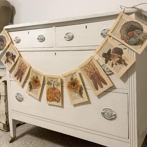 May include: A garland of vintage book pages with fall-themed illustrations, including a turkey, a pilgrim hat, corn, sunflowers, and a pumpkin. The garland is strung with twine and hangs in front of a white dresser.