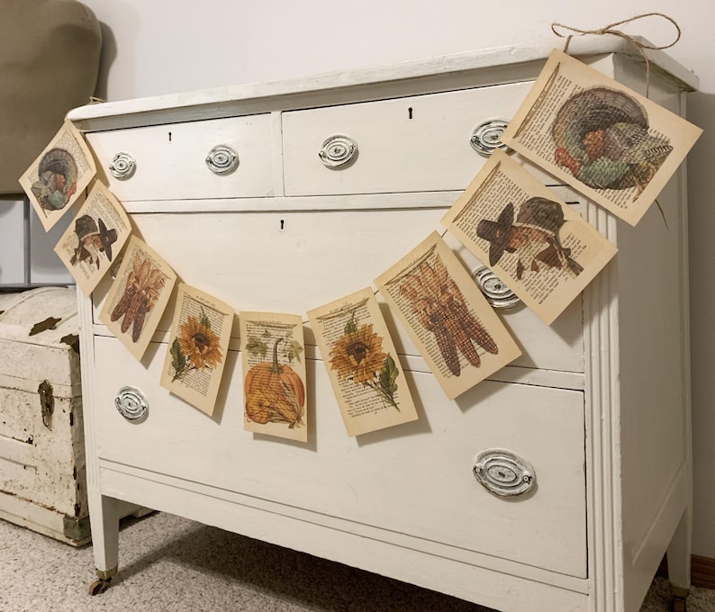May include: A garland of vintage book pages with fall-themed illustrations, including a turkey, a pilgrim hat, corn, sunflowers, and a pumpkin. The garland is strung with twine and hangs in front of a white dresser.