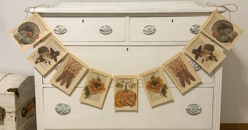 May include: A fall-themed banner with images of a turkey, a pilgrim, gloves, a sunflower, and a pumpkin. The banner is strung across a white dresser with silver hardware. The images are printed on pages from a book.