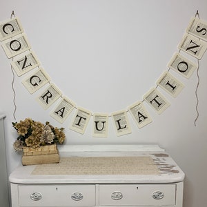 Pu&ograve; includere: Uno sfondo bianco con uno striscione che recita "CONGRATULATIONS" in lettere nere. Lo striscione &egrave; fatto di carta con un design di pagina di libro vintage. Lo striscione &egrave; appeso a un muro bianco con una corda. Un piccolo mazzo di fiori secchi &egrave; in una scatola di legno su una cassettiera bianca.