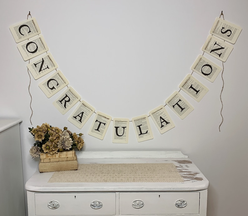 Pu&ograve; includere: Uno sfondo bianco con uno striscione che recita "CONGRATULATIONS" in lettere nere. Lo striscione &egrave; fatto di carta con un design di pagina di libro vintage. Lo striscione &egrave; appeso a un muro bianco con una corda. Un piccolo mazzo di fiori secchi &egrave; in una scatola di legno su una cassettiera bianca.