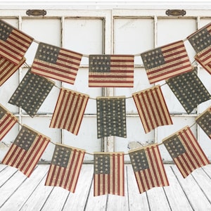 May include: A decorative bunting garland featuring multiple rectangular flags printed with the United States flag design. The flags are arranged in two cascading rows, suspended by a thin cord, and display a weathered, textured appearance.
