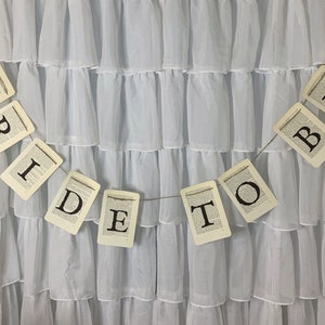 Pu&ograve; includere: Uno sfondo in tessuto bianco con uno striscione che recita "Bride to Be" in lettere nere su sfondo bianco. Lo striscione &egrave; composto da singole carte con ogni lettera stampata su una carta separata.