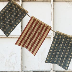 May include: A decorative bunting banner featuring three rectangular pennants suspended on a thin twine string. Two pennants display a dark blue background with a repeating pattern of light-colored stars, while the center pennant features red stripes.
