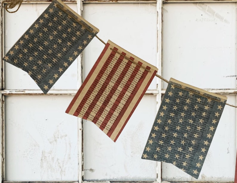 May include: A decorative bunting banner featuring three rectangular pennants suspended on a thin twine string. Two pennants display a dark blue background with a repeating pattern of light-colored stars, while the center pennant features red stripes.