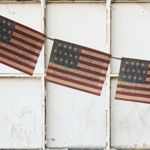 May include: A decorative bunting featuring three rectangular flags suspended on a thin cord. Each flag displays the American flag design with red and beige stripes and a blue canton containing white stars, all printed over a vintage book page.