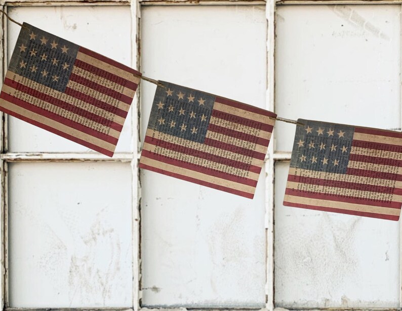 May include: A decorative bunting featuring three rectangular flags suspended on a thin cord. Each flag displays the American flag design with red and beige stripes and a blue canton containing white stars, all printed over a vintage book page.