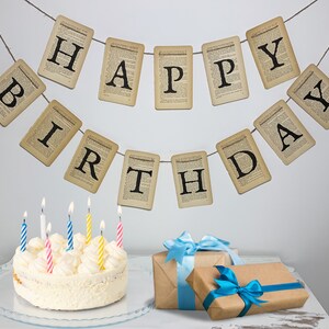 Pu&ograve; includere: Una torta bianca con glassa bianca e candele colorate &egrave; su un piatto da torta trasparente. Un banner con le parole "Buon compleanno" &egrave; appeso a un muro bianco dietro la torta.