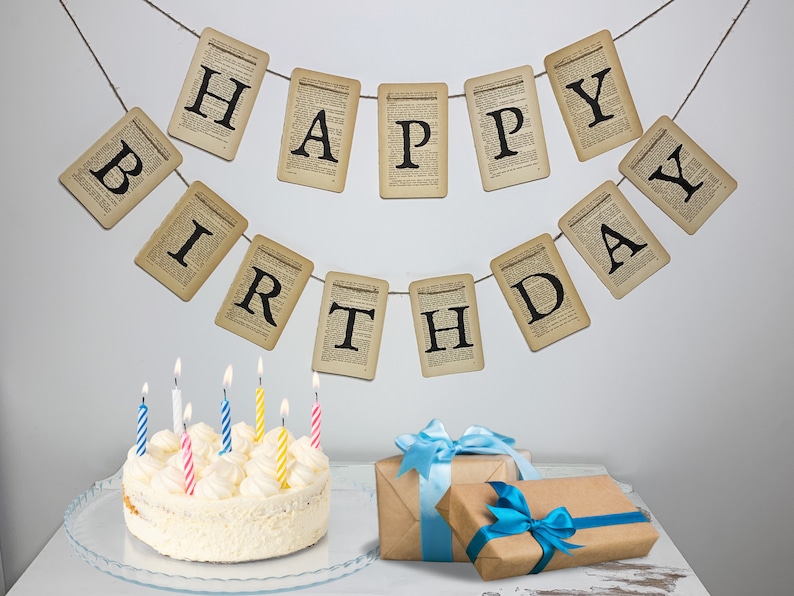 Pu&ograve; includere: Una torta bianca con glassa bianca e candele colorate &egrave; su un piatto da torta trasparente. Un banner con le parole "Buon compleanno" &egrave; appeso a un muro bianco dietro la torta.