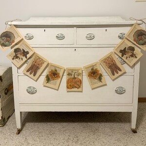 May include: A garland of vintage-style paper cards with fall-themed illustrations, including pumpkins, sunflowers, and turkeys, hangs in front of a white dresser. The cards are strung together with twine, creating a rustic, seasonal decoration.