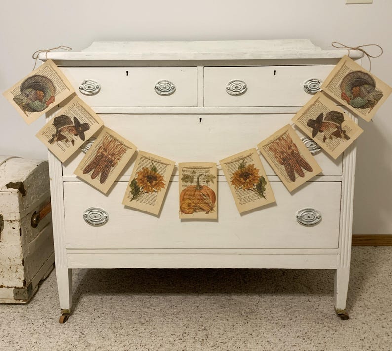 May include: A garland of vintage-style paper cards with fall-themed illustrations, including pumpkins, sunflowers, and turkeys, hangs in front of a white dresser. The cards are strung together with twine, creating a rustic, seasonal decoration.