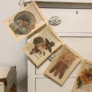 May include: Thanksgiving-themed banner featuring illustrations of a turkey, pilgrim, corn, and sunflower. Each image is printed on a page from a vintage book, creating a rustic and charming aesthetic. The banner is strung together with twine.
