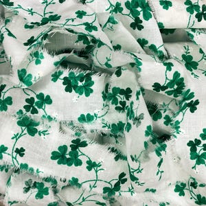 May include: White fabric with a repeating pattern of green shamrocks and vines. The edges of the fabric are frayed, giving it a textured appearance. The shamrocks are a vibrant green, and the fabric appears to be lightweight.
