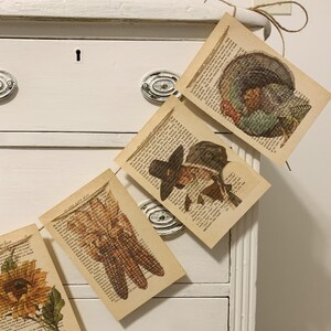 May include: Thanksgiving-themed garland featuring vintage book pages with images of a turkey, corn, and a pilgrim. The garland is strung on twine and hangs against a white dresser.