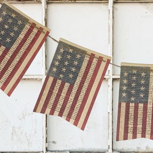 May include: A decorative bunting banner featuring rectangular flags printed with the American flag design. Each flag has a blue canton with white stars and red and white stripes, with the entire surface overlaid with vintage-style printed text.